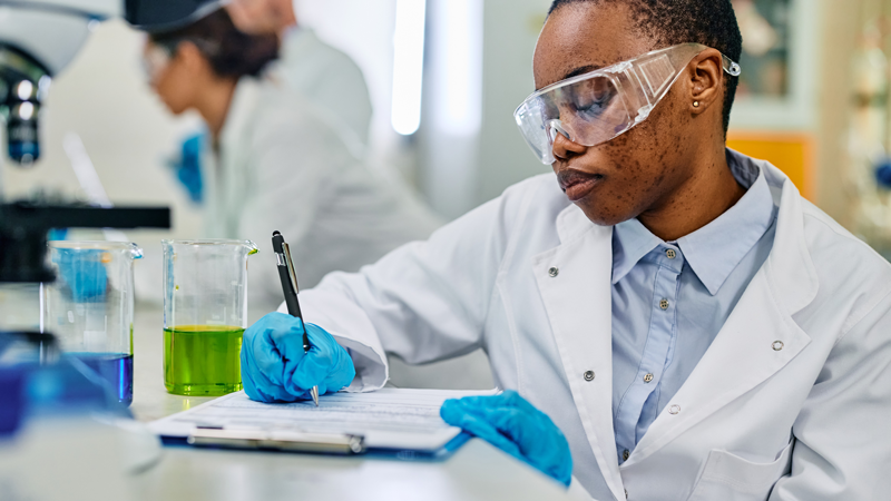 A young Black scientist wearing a lab coat, safety goggles, and gloves writes research data on a clipboard in a laboratory